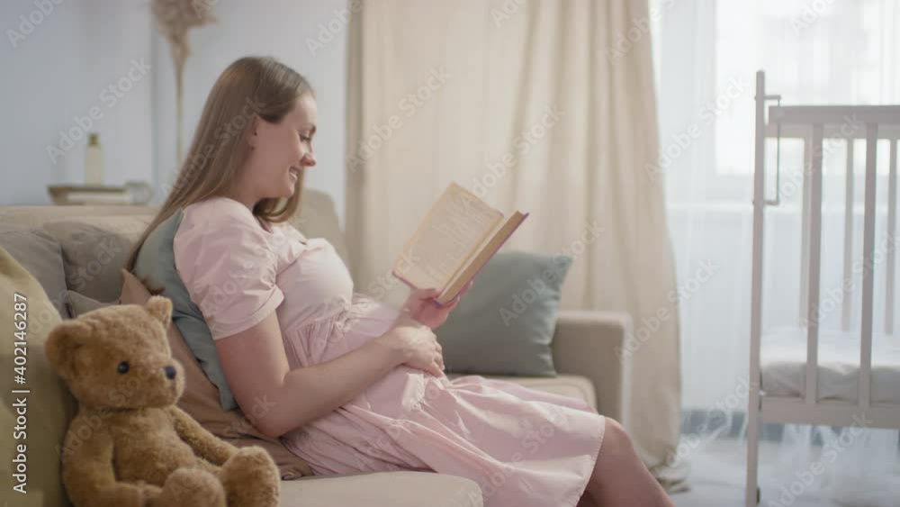 Medium slow-motion side-view of smiling pregnant woman sitting on sofa in living room next to teddy bear holding book reading aloud while stroking belly Medium slow-motion side-view of smiling pregnant woman sitting on sofa in living room next to teddy bear holding book reading aloud while stroking belly