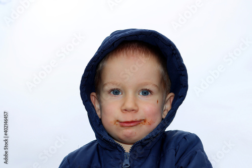 Portrait of a three-year-old boy with a chocolate-stained face on a white background. Boy with a dirty face and a blue jacket with a hood.