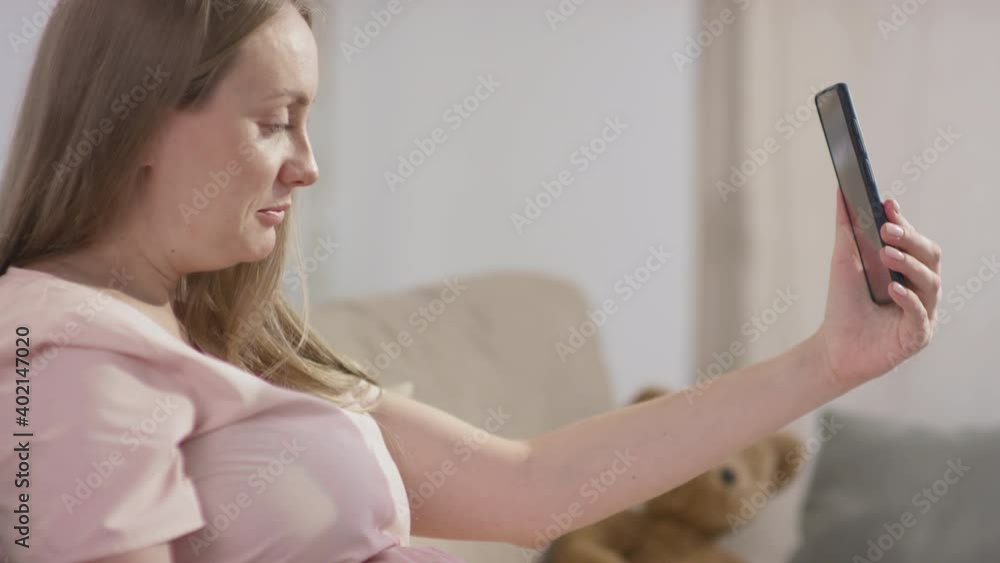 Tilting slow-motion side-view medium close-up of young smiling pregnant woman having video call sitting in living room alone Tilting slow-motion side-view medium close-up of young smiling pregnant woman having video call sitting in living room alone