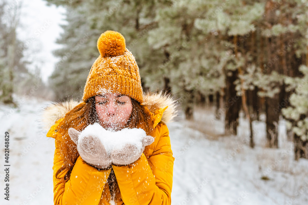 Outdoor close-up portrait of a young stylish smiling smiling girl wearing a yellow jacket and a knitted hat, walking in the winter forest and playing with the snow outdoors. Christmas holidays