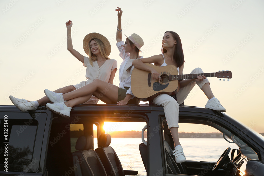 Happy friends with guitar sitting on car roof outdoors at sunset. Summer trip Happy friends with guitar sitting on car roof outdoors at sunset. Summer trip