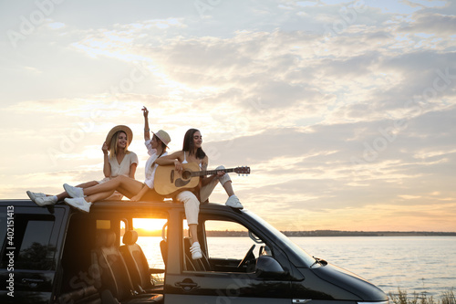 Happy friends with guitar sitting on car roof outdoors at sunset. Summer trip © New Africa