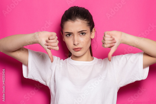 Dissatisfied displeased disgusted young brunette woman 20s wearing casual basic shirt standing showing thumb down looking camera isolated on pastel pink colour background, studio portrait © Misha