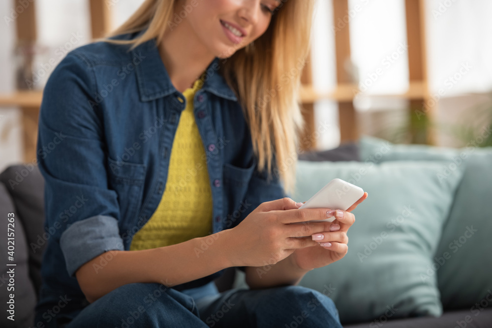 Cropped view of blonde woman texting on smartphone while sitting on couch on blurred background