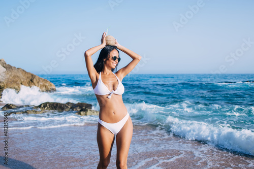 Happy female tourist with slim figure smiling at coastline beach enjoying sunbathing time during sunny day, cheerful sexy girl with coconut beverage rejoicing at seashore on Philippines island © BullRun