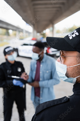 Police officer in medical mask and sunglasses standing near african american victim and colleague on blurred background.
