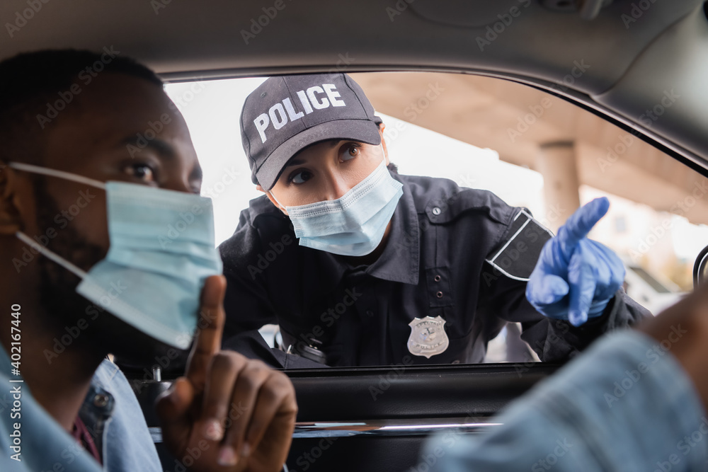 Policewoman in medical mask and latex glove pointing with finger near african american driver sitting in car on blurred foreground. Policewoman in medical mask and latex glove pointing with finger near african american driver sitting in car on blurred foreground.