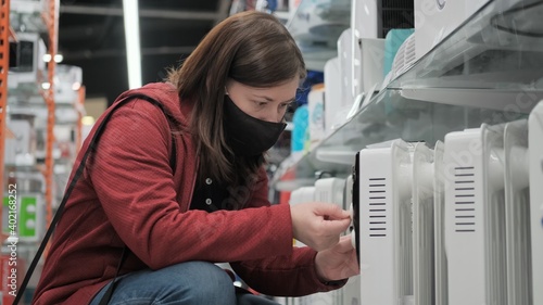 Women wearing antivirus medical mask picks out portable heater in electronics shop. Going to showroom during pandemic. Screen from video footage photo