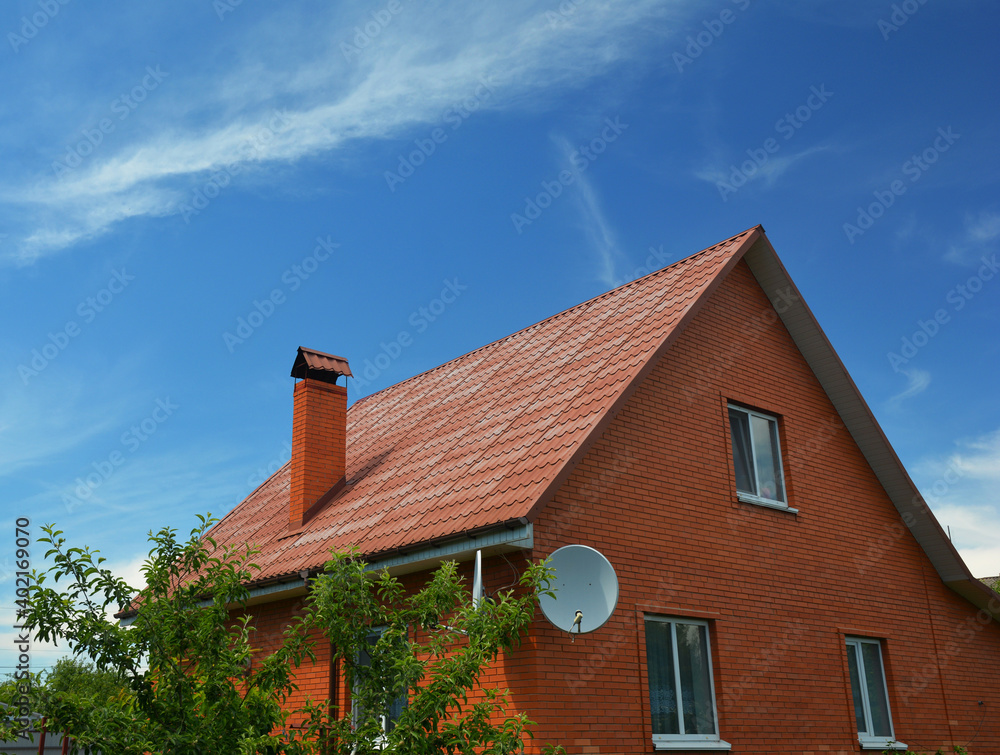 A house of simple construction built from red bricks with a metal gabled roof, a chimney, roof gutters, an attic window, and a satellite dish antenna against the blue sky. A house of simple construction built from red bricks with a metal gabled roof, a chimney, roof gutters, an attic window, and a satellite dish antenna against the blue sky.