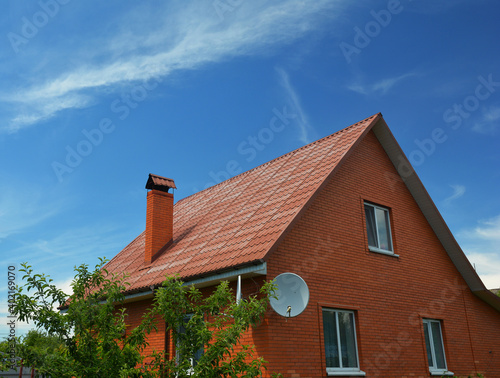 A house of simple construction built from red bricks with a metal gabled roof, a chimney, roof gutters, an attic window, and a satellite dish antenna against the blue sky.