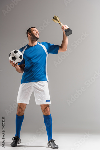 Fototapeta Naklejka Na Ścianę i Meble -  Smiling sportsman holding football and looking at champions trophy on grey background