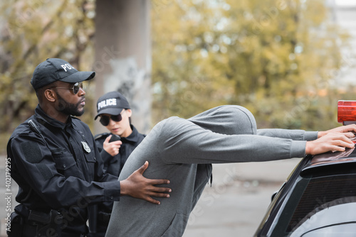 african american policeman frisking hooded offender leaning on patrol car near colleague on blurred background outdoors.