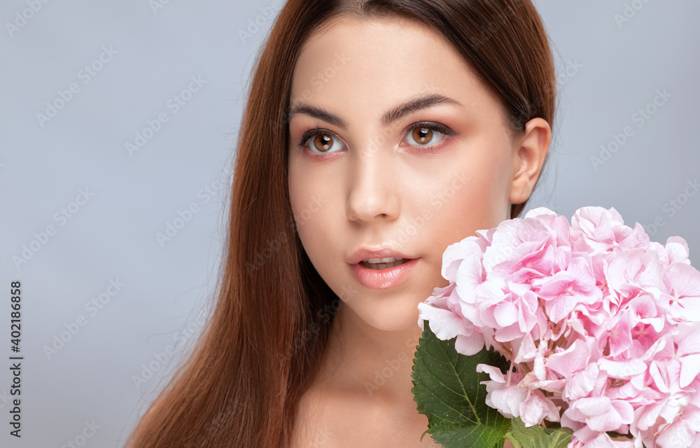 Portrait of a beautiful brunette woman with healthy clean skin and fresh make-up. She holds a pink hydrangea near her face. Aesthetic cosmetology and makeup concept.