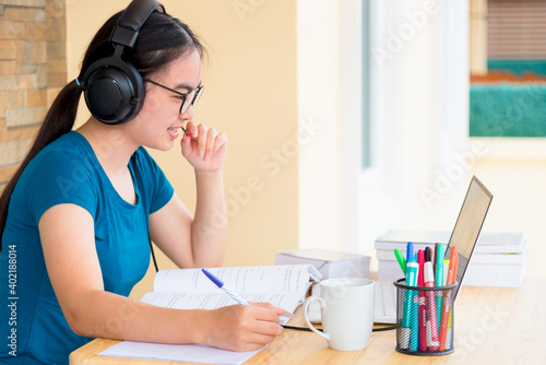 Asian teenage girl student with a headset and eyeglasses looks at laptop computer online learning from school and speak into a microphone. Distance education class of the college video call at a home