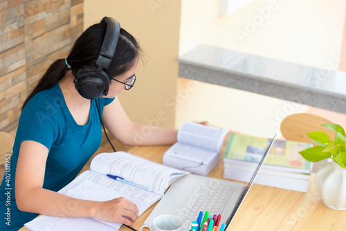 Young Asian teenage girl student with a headset glasses sitting on the desk reading a book using laptop computer online study from school. Education from the class of university by video call at home