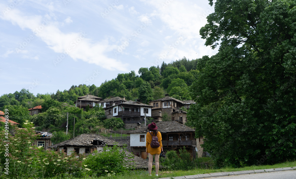 Girl Backpacker alone visiting old village on a trip in Bulgaria. Caucasian woman with hat standing searching and looking in front of mountain town. Eastern balkan calm rural destination
