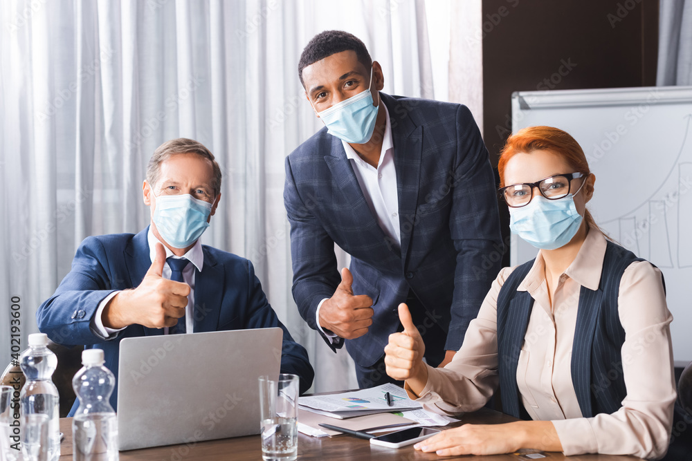 Multicultural businesspeople in medical masks looking at camera while showing thumbs up at workplace.