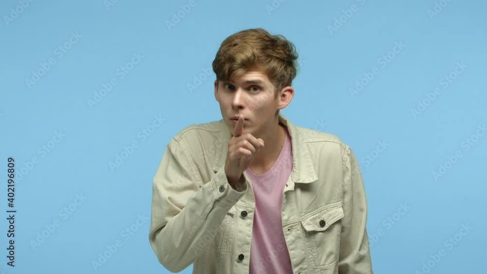 Slow motion of young angry guy shushing at camera, hushing and pressing finger to lips, pointing at camera, telling to shut mouth and be quiet, standing over blue background Slow motion of young angry guy shushing at camera, hushing and pressing finger to lips, pointing at camera, telling to shut mouth and be quiet, standing over blue background