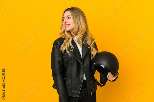 Young blonde caucasian biker woman holding helmet looks aside smiling, cheerful and pleasant.