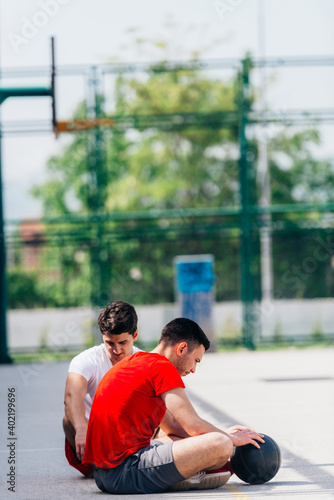 Two strong caucasian athletes resting on the ground at the basketball court while having a conversation.
