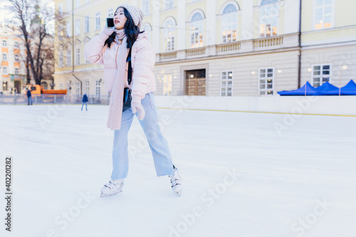 Full length portrait of a young asian female wearing powder pink jacket and warm knitted hat, scarf and gloves taking on ice rink talking on the phone, outdoors at winter time. photo