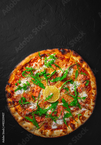 Pizza with Mozzarella cheese, salmon fish, tomato sauce, arugula, lemon. Italian pizza on Dark grey black slate background