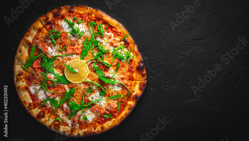 Pizza with Mozzarella cheese, salmon fish, tomato sauce, arugula, lemon. Italian pizza on Dark grey black slate background