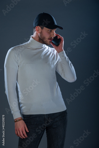 Male model with a black hat and black mobile phone posing in a studio wearing white blouse and black pants