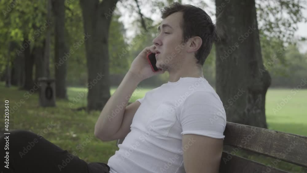 Side view of young millennial brunette man sitting on bench on summer park alley talking on the phone. Carefree relaxed Caucasian guy resting outdoors. Lifestyle and leisure concept. Side view of young millennial brunette man sitting on bench on summer park alley talking on the phone. Carefree relaxed Caucasian guy resting outdoors. Lifestyle and leisure concept.