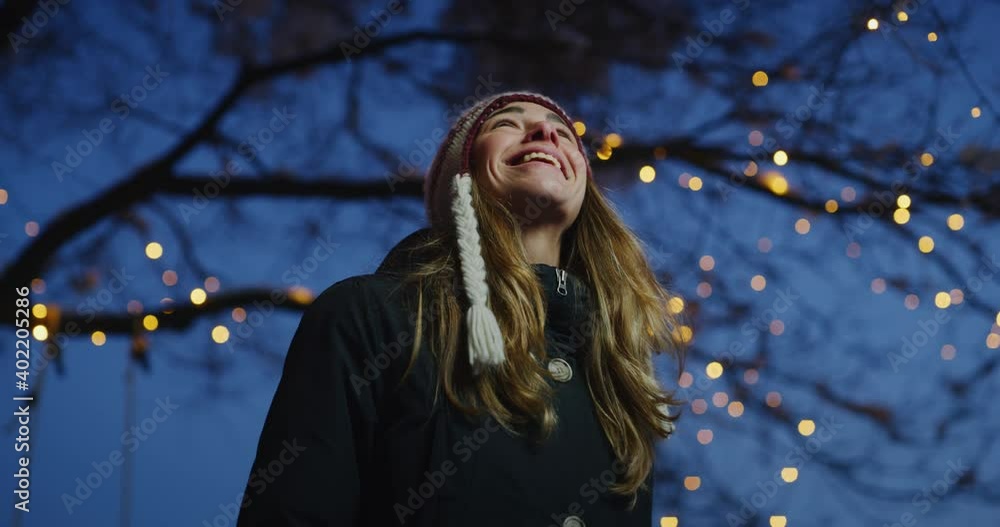 Cinematic shot of carefree happy smiling young woman wearing warm hat is excited and amazed with Christmas lights on the tree in a festive winter evening.Concept of winter holidays, freedom, happiness Cinematic shot of carefree happy smiling young woman wearing warm hat is excited and amazed with Christmas lights on the tree in a festive winter evening.Concept of winter holidays, freedom, happiness
