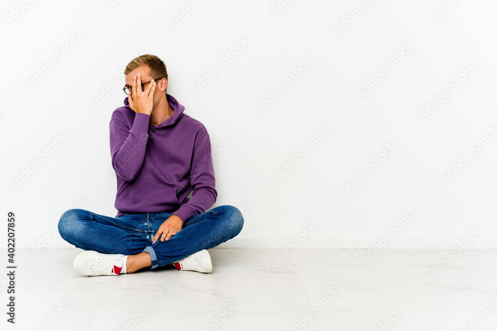 Young caucasian man sitting on the floor blink at the camera through fingers, embarrassed covering face. Young caucasian man sitting on the floor blink at the camera through fingers, embarrassed covering face.