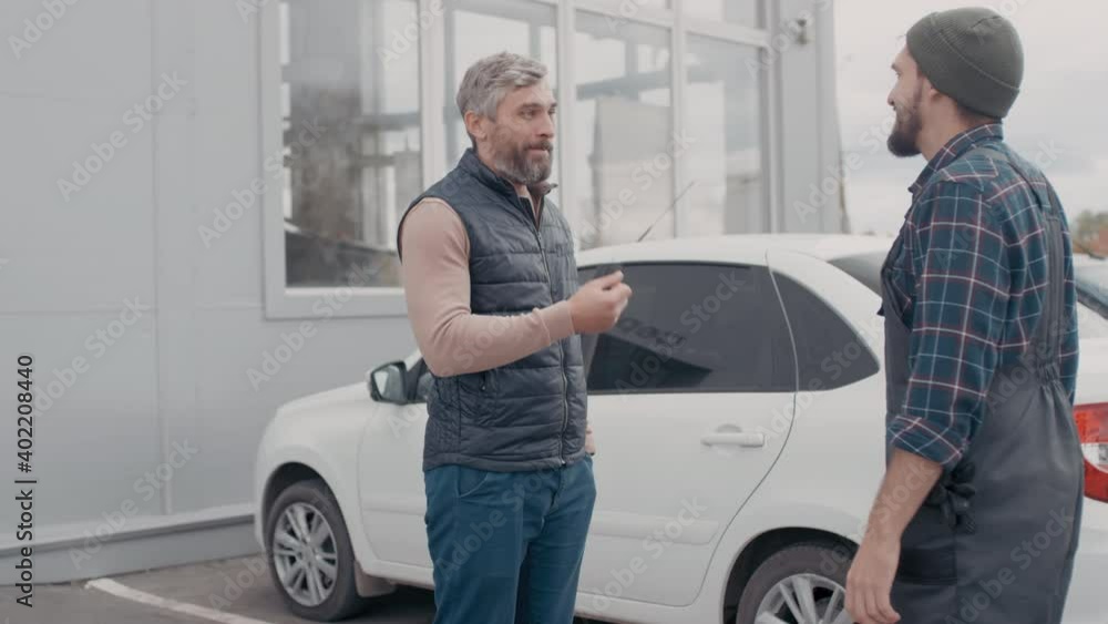 Handheld shot of bearded male auto mechanic in overalls talking to client and returning his car keys after finishing repairs Handheld shot of bearded male auto mechanic in overalls talking to client and returning his car keys after finishing repairs