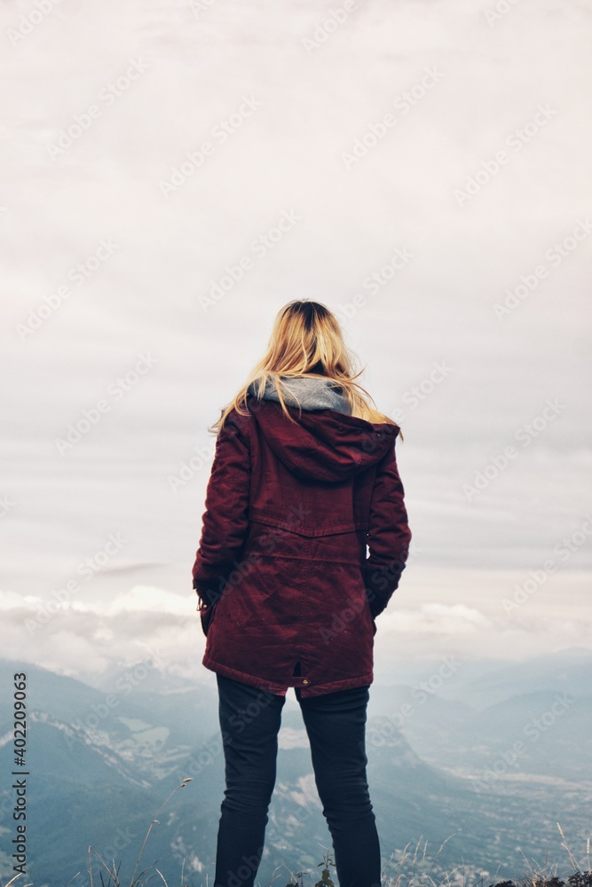 Young blond woman facing away standing on a cliff in winter wearing dark red burgundy coat looking into the distance to the foggy mountains Young blond woman facing away standing on a cliff in winter wearing dark red burgundy coat looking into the distance to the foggy mountains