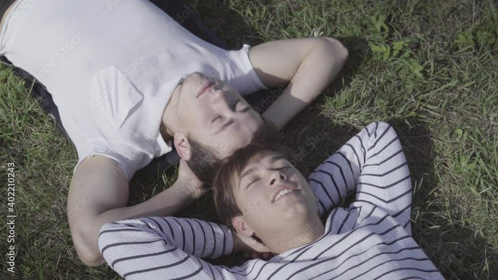 Close-up top view of relaxed young men lying on green summer grass with closed eyes. Portrait of carefree friends or lovers resting outdoors in sunlight. Friendship or relationship concept. Close-up top view of relaxed young men lying on green summer grass with closed eyes. Portrait of carefree friends or lovers resting outdoors in sunlight. Friendship or relationship concept.