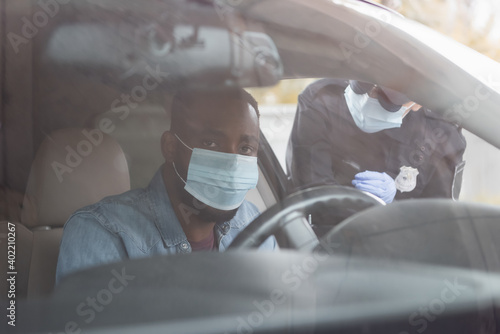 African american driver in medical mask sitting in car near policewoman on blurred background.