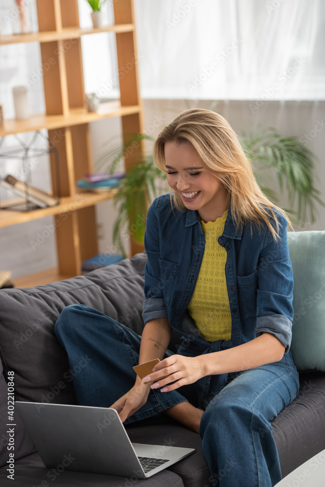 blonde woman with credit card laughing while sitting on couch near laptop on blurred background blonde woman with credit card laughing while sitting on couch near laptop on blurred background