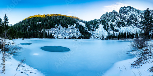 A frozen mountain lake surrounded by snow, forests and cliffs with a clear blue sky