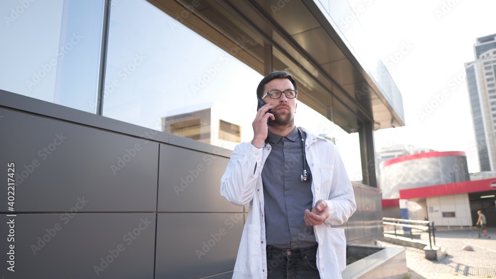 Fototapeta premium MS doctor in white coat talking on mobile phone front of clinic. Man medic outside speaking on cell phone. Medicine healthcare professional theme. Physician discussing with patient during phone talk