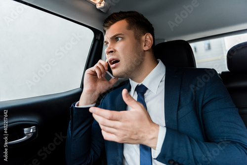 Angry businessman talking on smartphone on back seat of car.