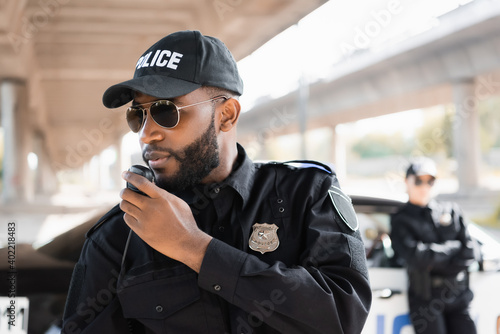 Fototapeta african american police officer talking on radio set near policewoman on blurred background outdoors