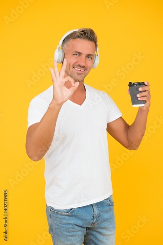 Happy man listen to music showing OK hand gesture for takeaway coffee in disposable cup yellow background, coffee-to-go © be free