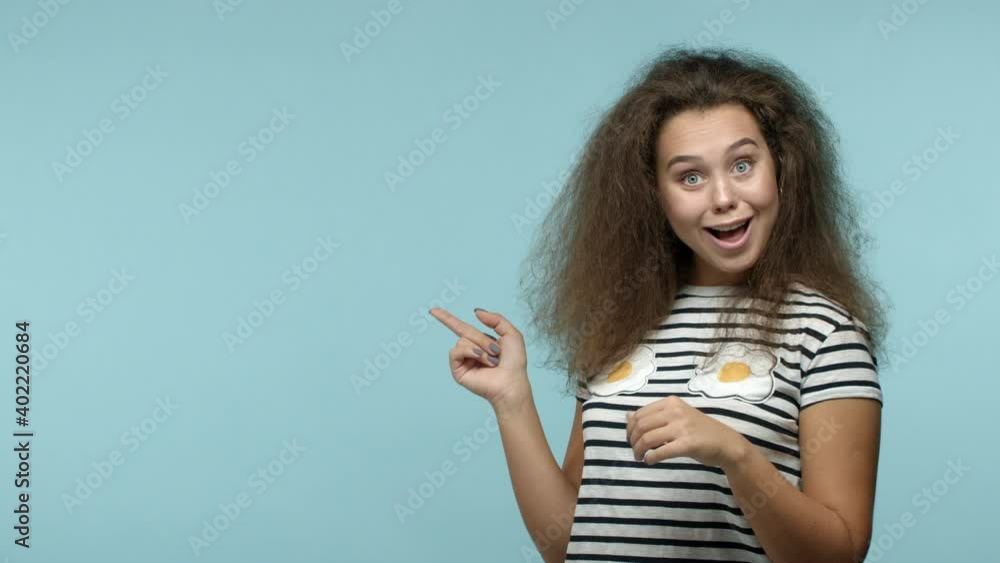Slow motion of silly cute girl in t-shirt turn head, looking and pointing left at copy space with your logo, showing thumbs up and smiling pleased, recommend company, blue background