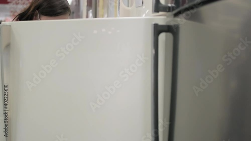 Woman in protective mask against virus chooses fridge made in vintage, retro style in home appliance store. Concept of shopping during pandemic
