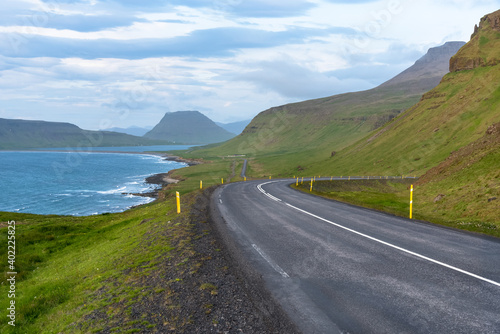 Empty winding coastal road running at the foot of grassy mountains in Iceland on a cloudy summer evening.