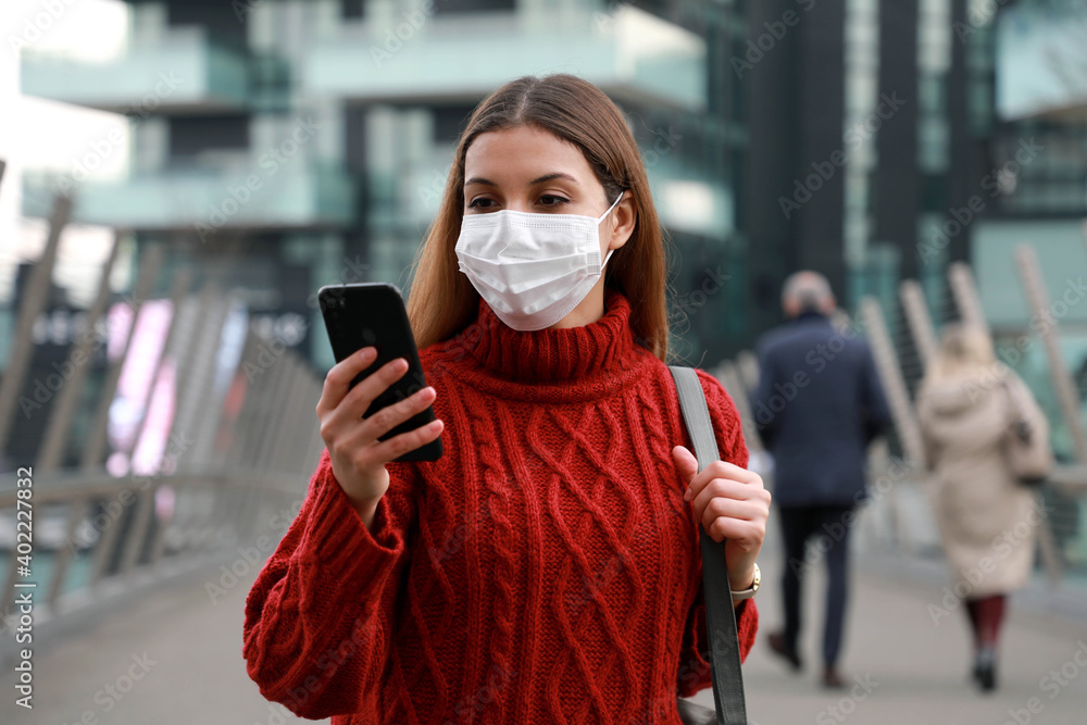 Fototapeta premium Young business woman walking along the city street wearing surgical mask and chatting with mobile phone. Office worker with protective mask using smartphone outdoor.