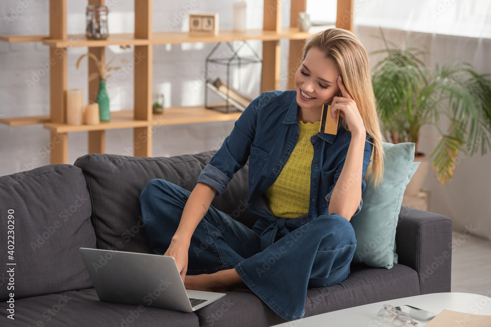 blonde woman with credit card using laptop while sitting on couch at home on blurred background blonde woman with credit card using laptop while sitting on couch at home on blurred background