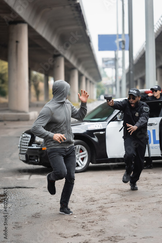 Φωτογραφία full length of hooded offender running from shocked african american policeman aiming with pistol on blurred background on urban street