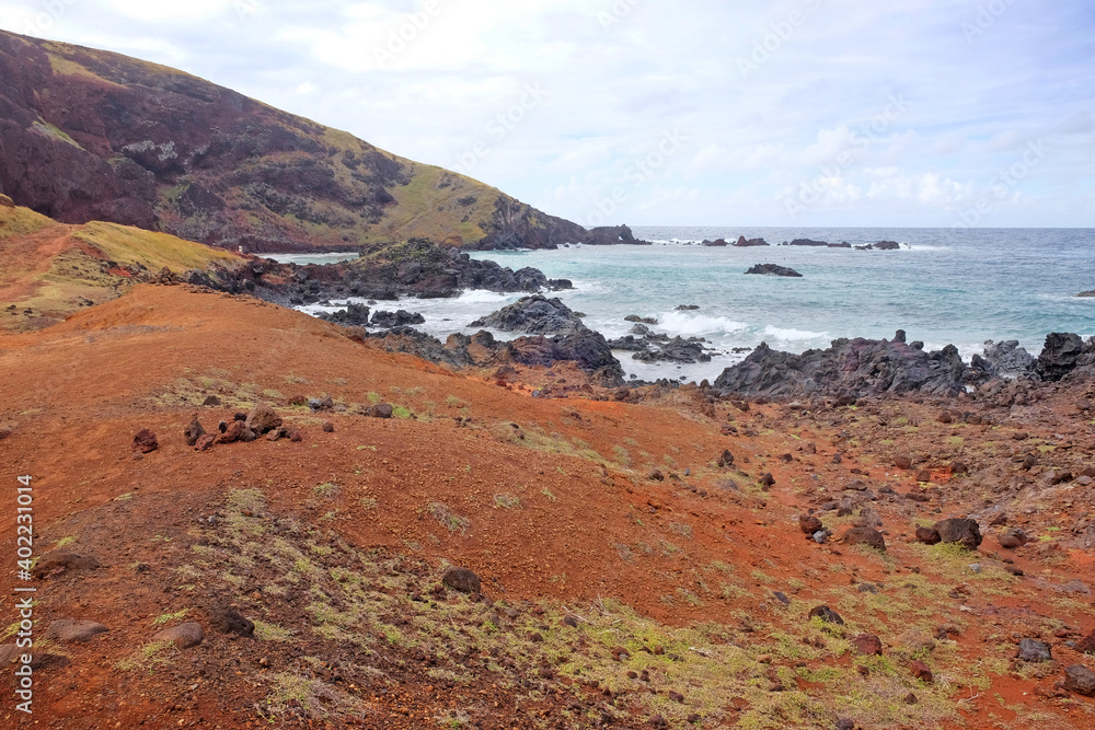 Small beach on the coast of Easter Island, covered by red volcanic dust, against a blue sky with white clouds.