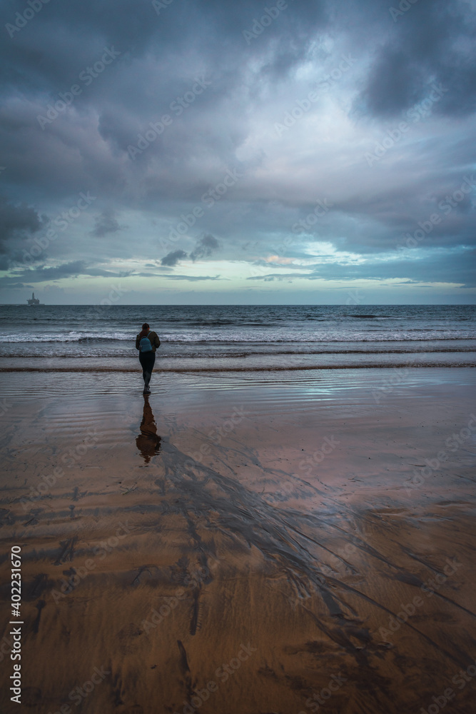 a girl with a blue backpack walking along a beach on a cloudy winter day