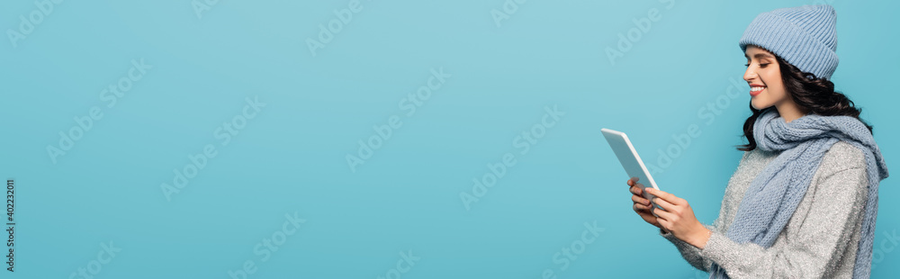 brunette woman in hat and scarf looking at digital tablet isolated on blue, banner brunette woman in hat and scarf looking at digital tablet isolated on blue, banner